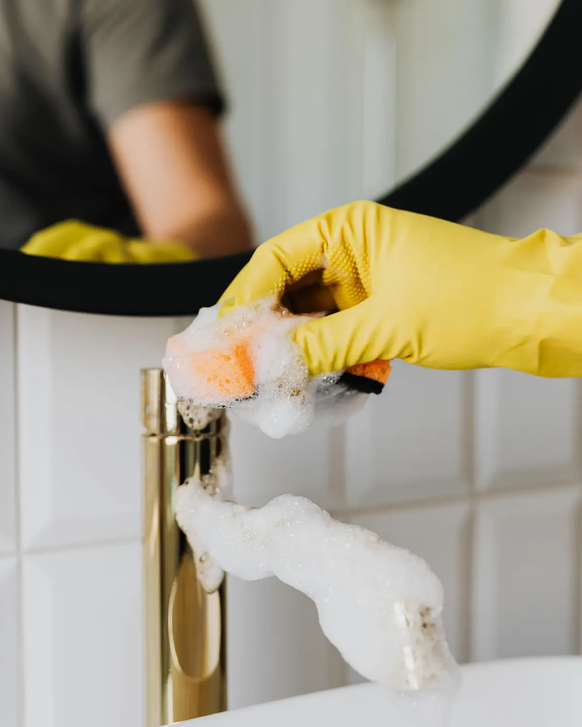 Gloved hand scrubbing a gold bathroom faucet with soapy foam