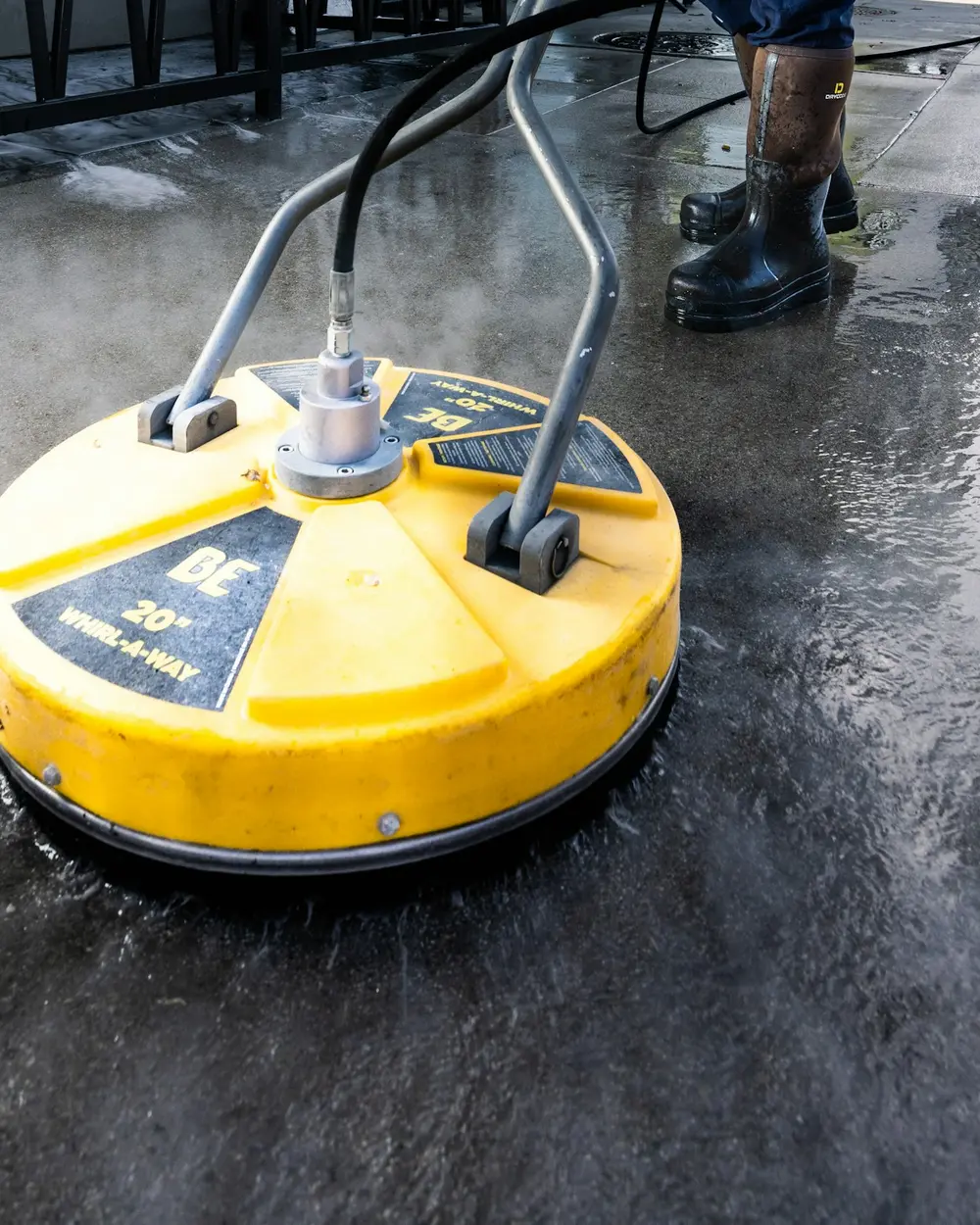 Worker in boots guiding a yellow surface cleaner across wet concrete