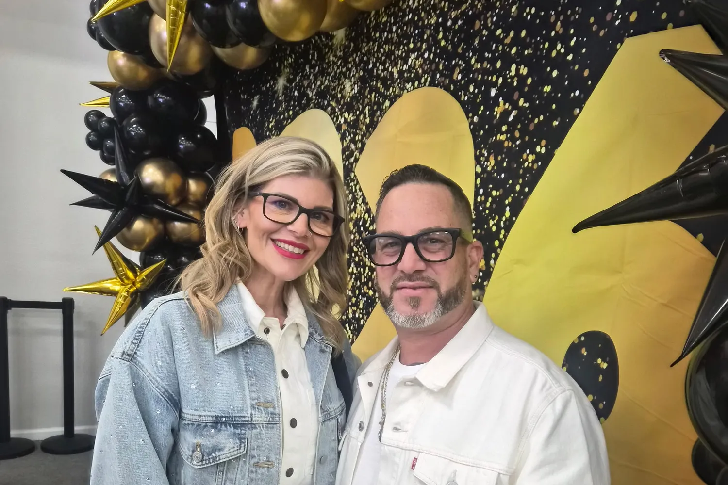 Lesley and Juni Martir, owners of Heavenly Handz, smiling together in front of black and gold balloons