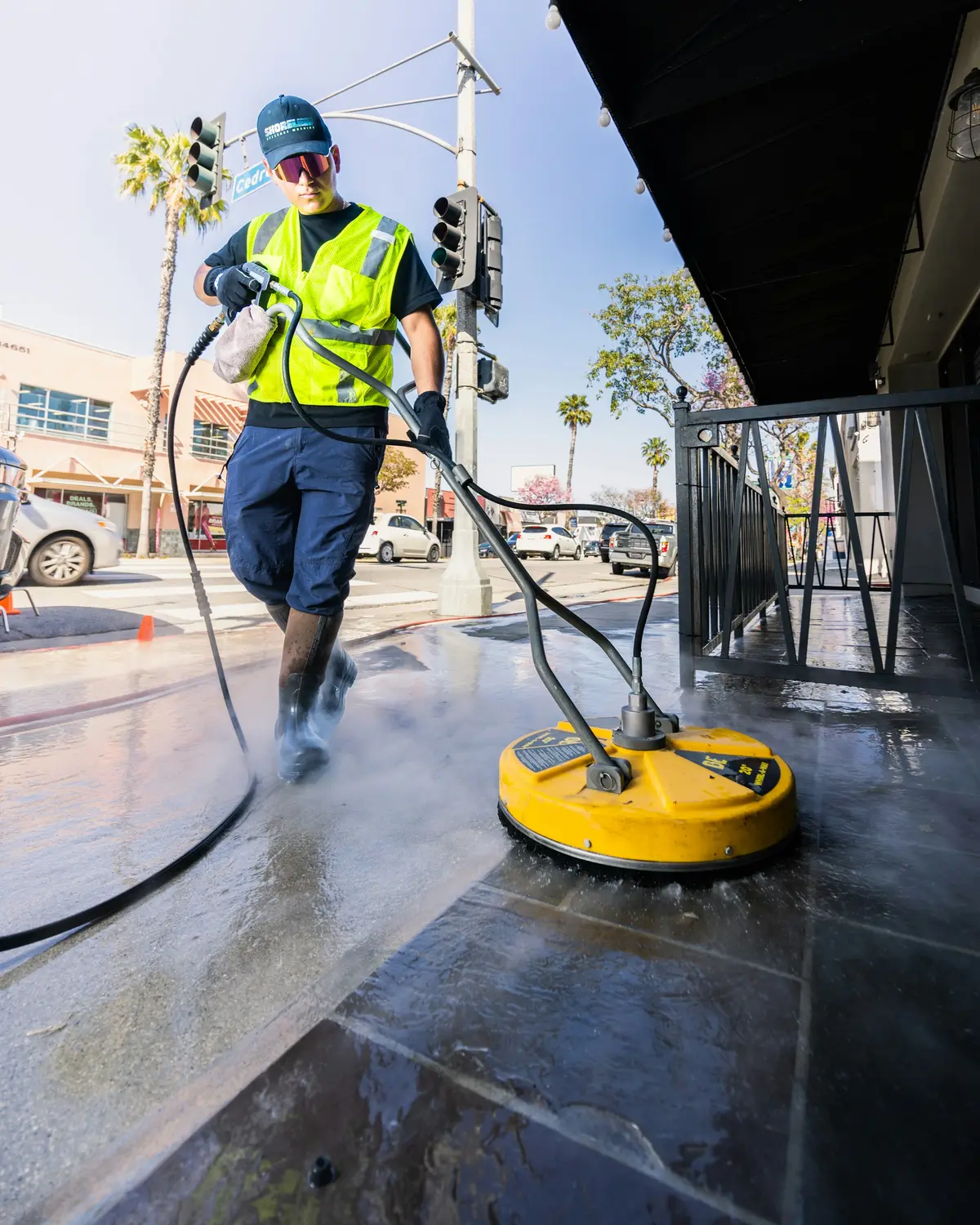 Worker running a surface cleaner across wet concrete beside palm trees
